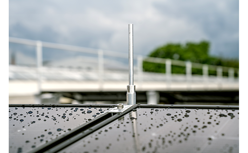 Close-up van een zonne-installatie met waterdruppels op het oppervlak en een gemonteerde bliksemafleider; op de achtergrond een reling voor valbeveiliging.